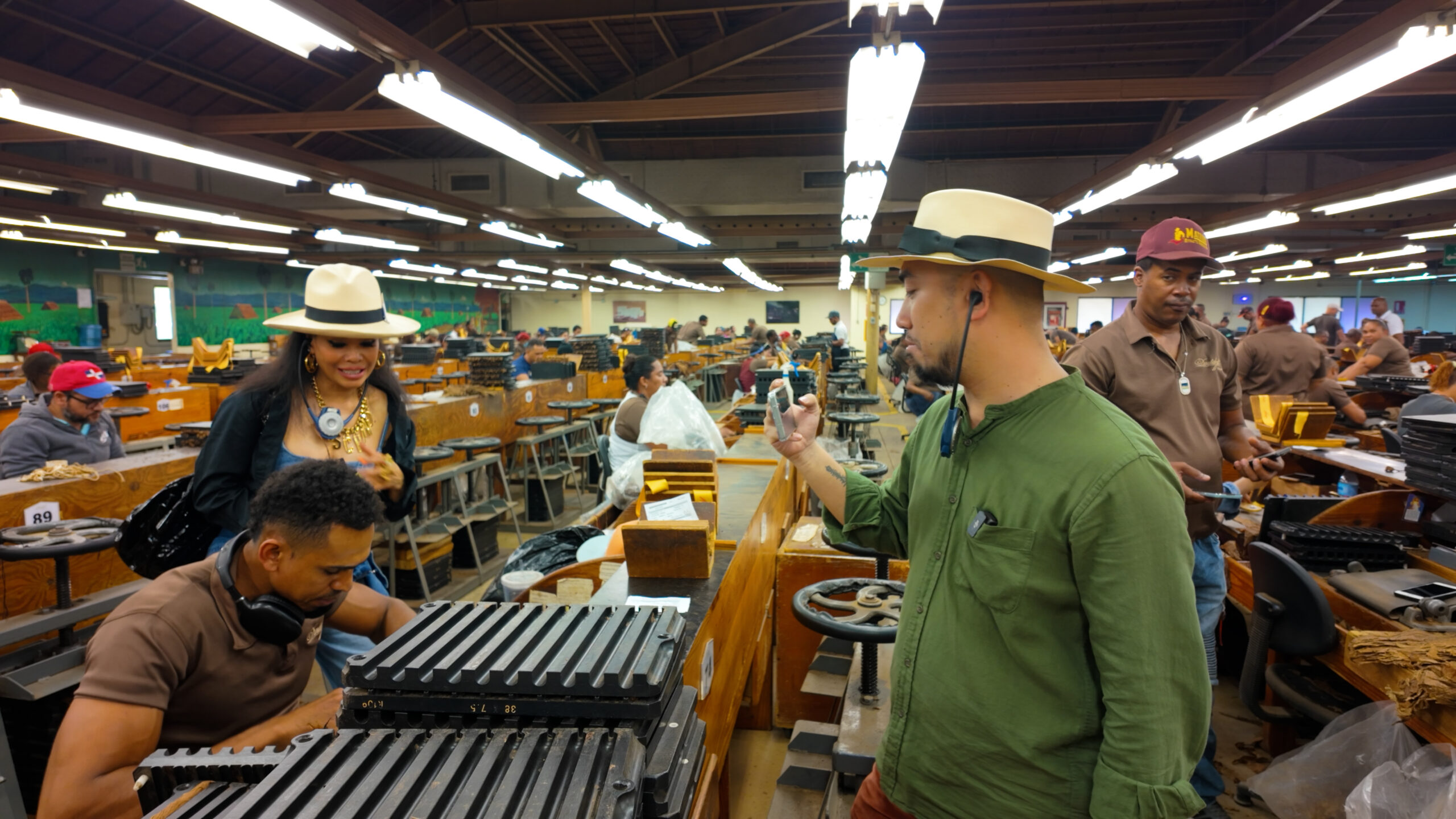 Nhà máy sản xuất xì gà với người lao động và khách tham quan đội mũ, trong không gian sáng đèn.