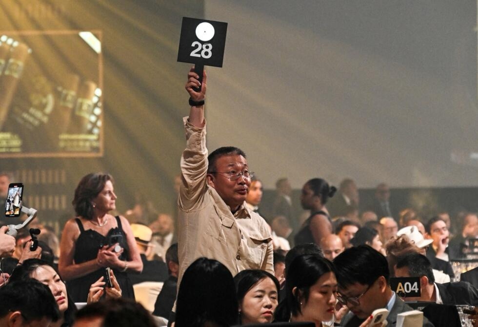 4-1-1 Người đàn ông giơ bảng số 28 tại phiên đấu giá đông người, nền sáng mờ và không khí sôi nổi.