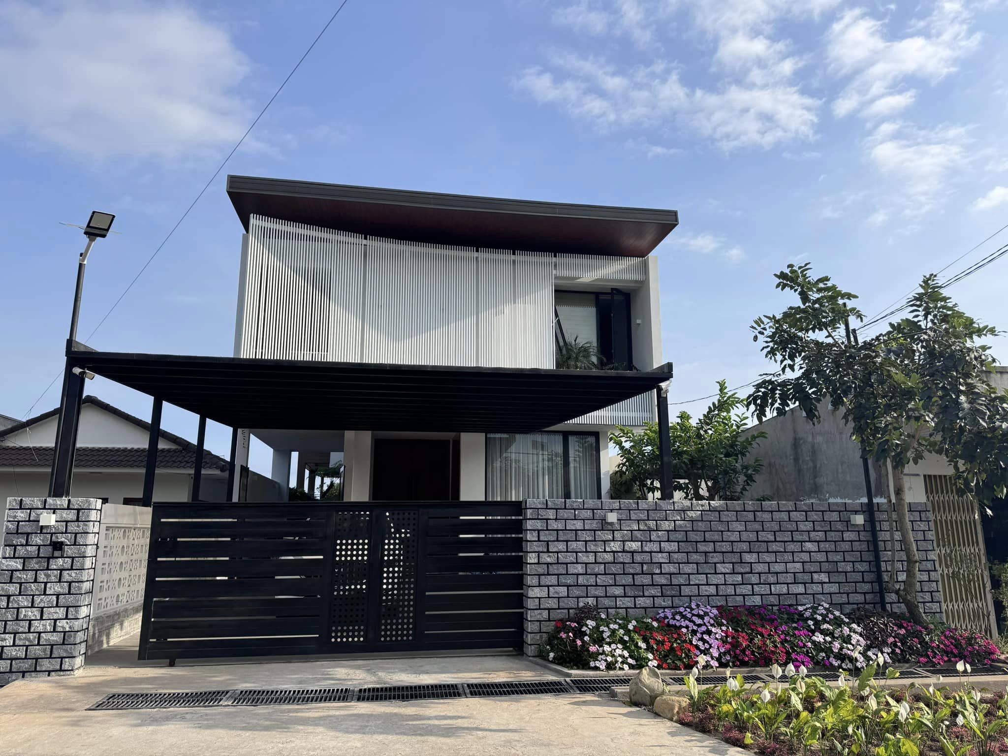 Ngôi nhà hiện đại hai tầng với mái hiên rộng và vườn hoa phía trước dưới bầu trời xanh.