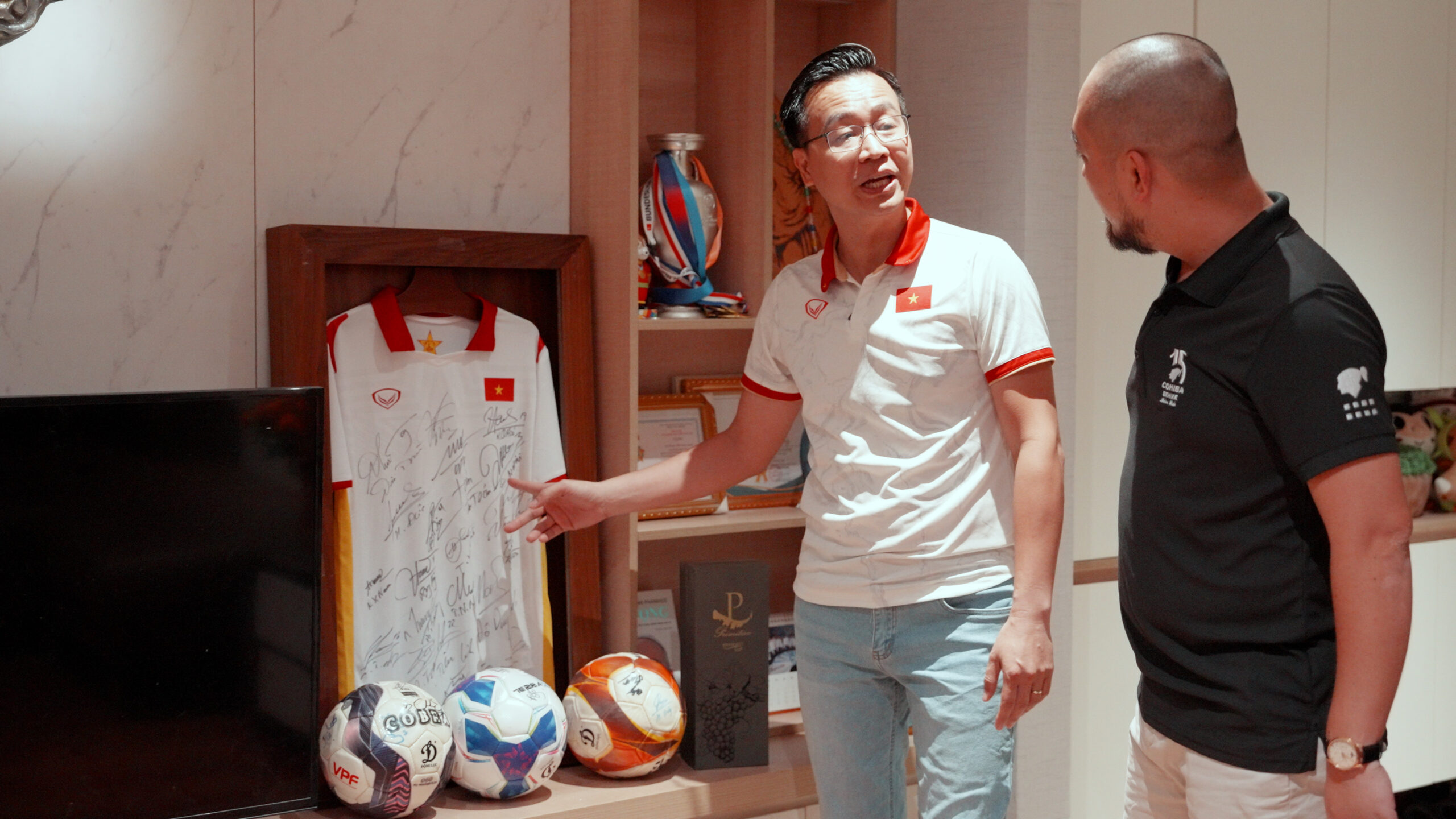 Hai người đàn ông nói chuyện cạnh áo bóng đá có chữ ký trong một phòng trưng bày.