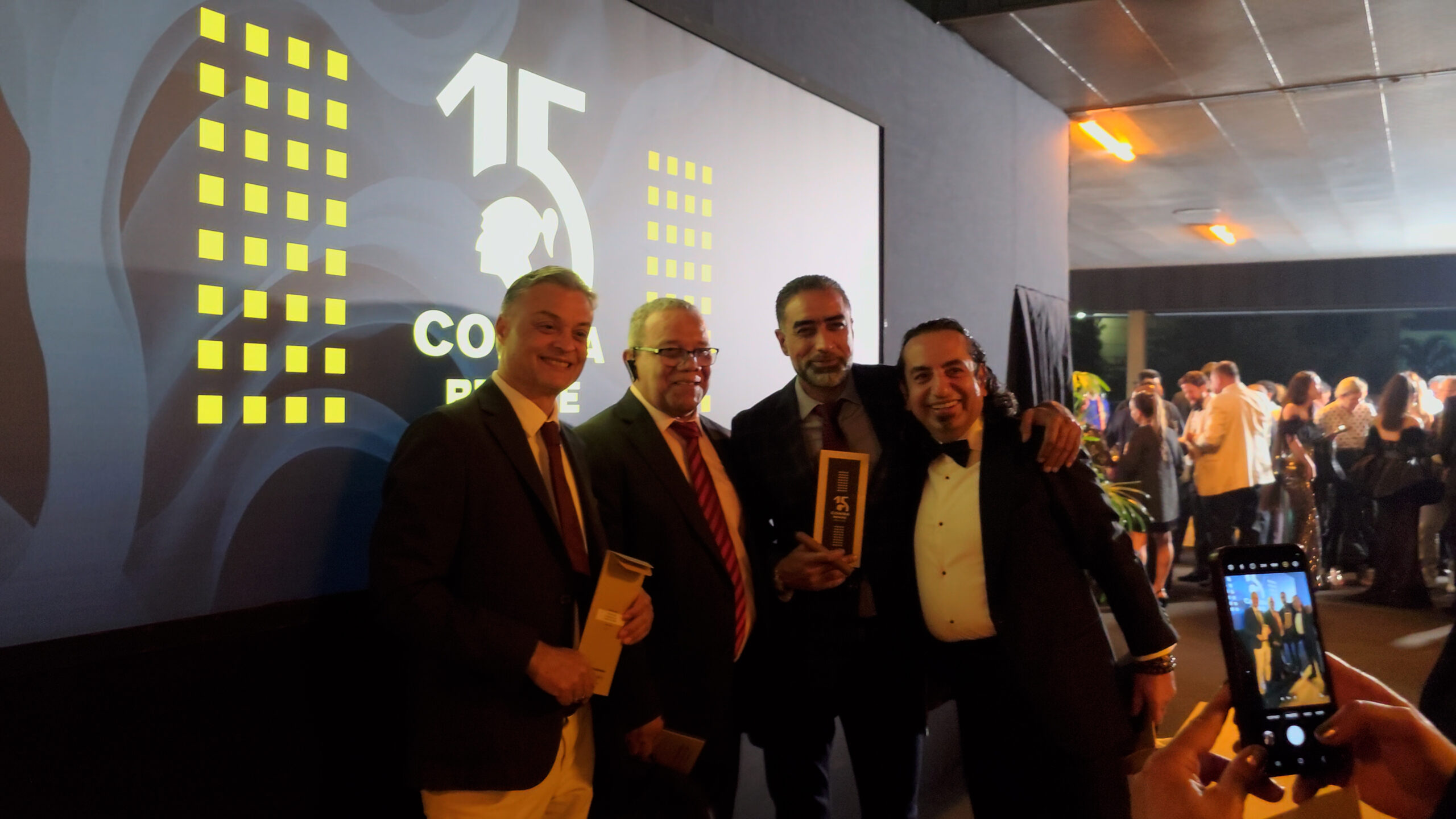 43-scaled-1 Bốn người đàn ông mặc vest đứng trước màn hình lớn tại sự kiện, một người đang chụp ảnh bằng điện thoại.