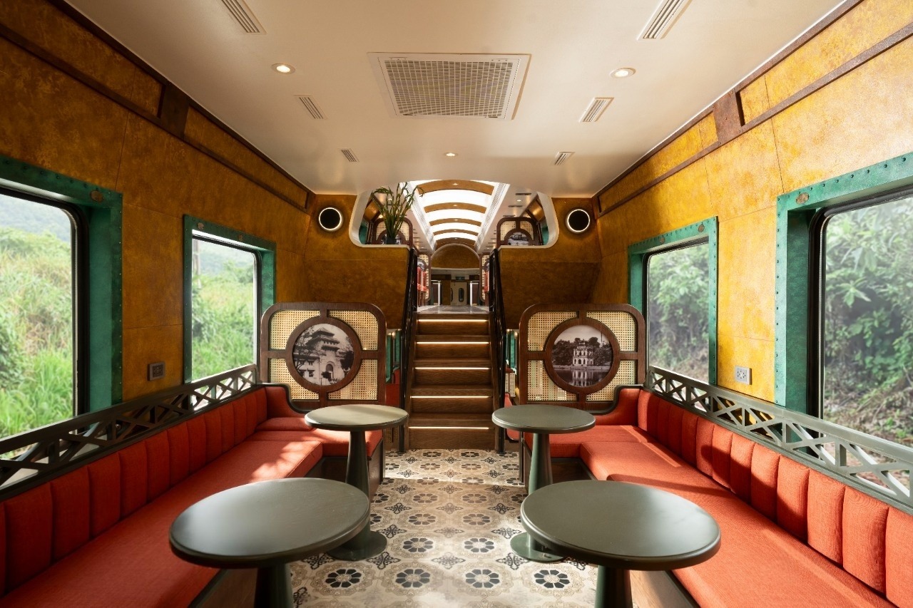 Nội thất toa tàu cổ điển với ghế bọc đỏ và bàn tròn, ánh sáng tự nhiên từ cửa sổ lớn hai bên.