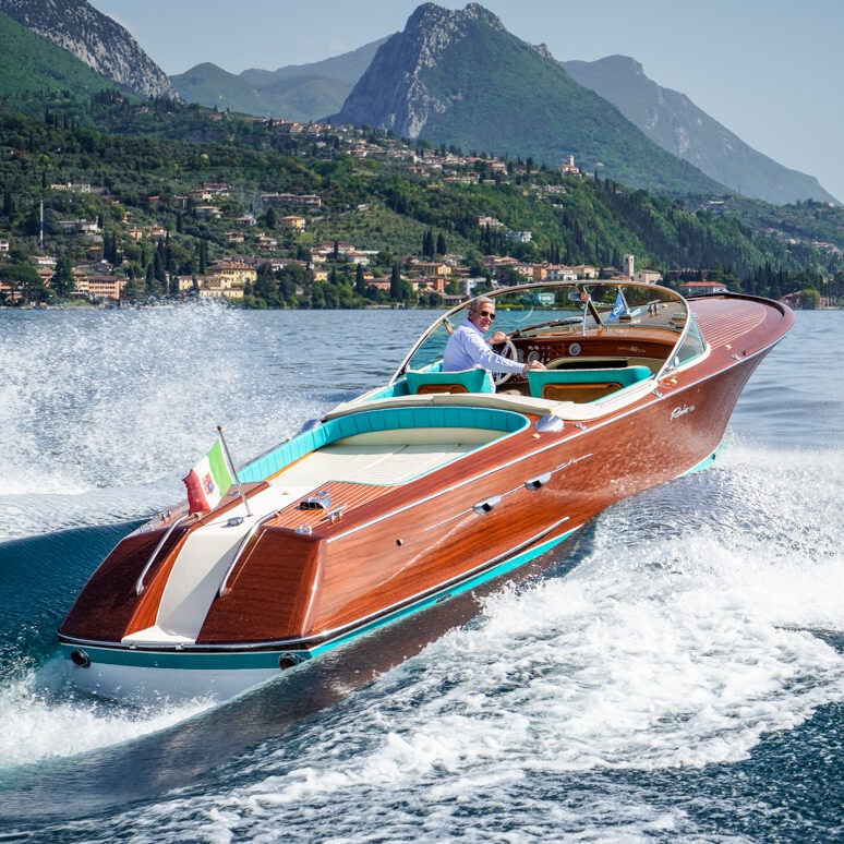 Thuyền gỗ sang trọng chạy trên mặt hồ xanh, có núi non bao quanh và lá cờ Ý phía sau.