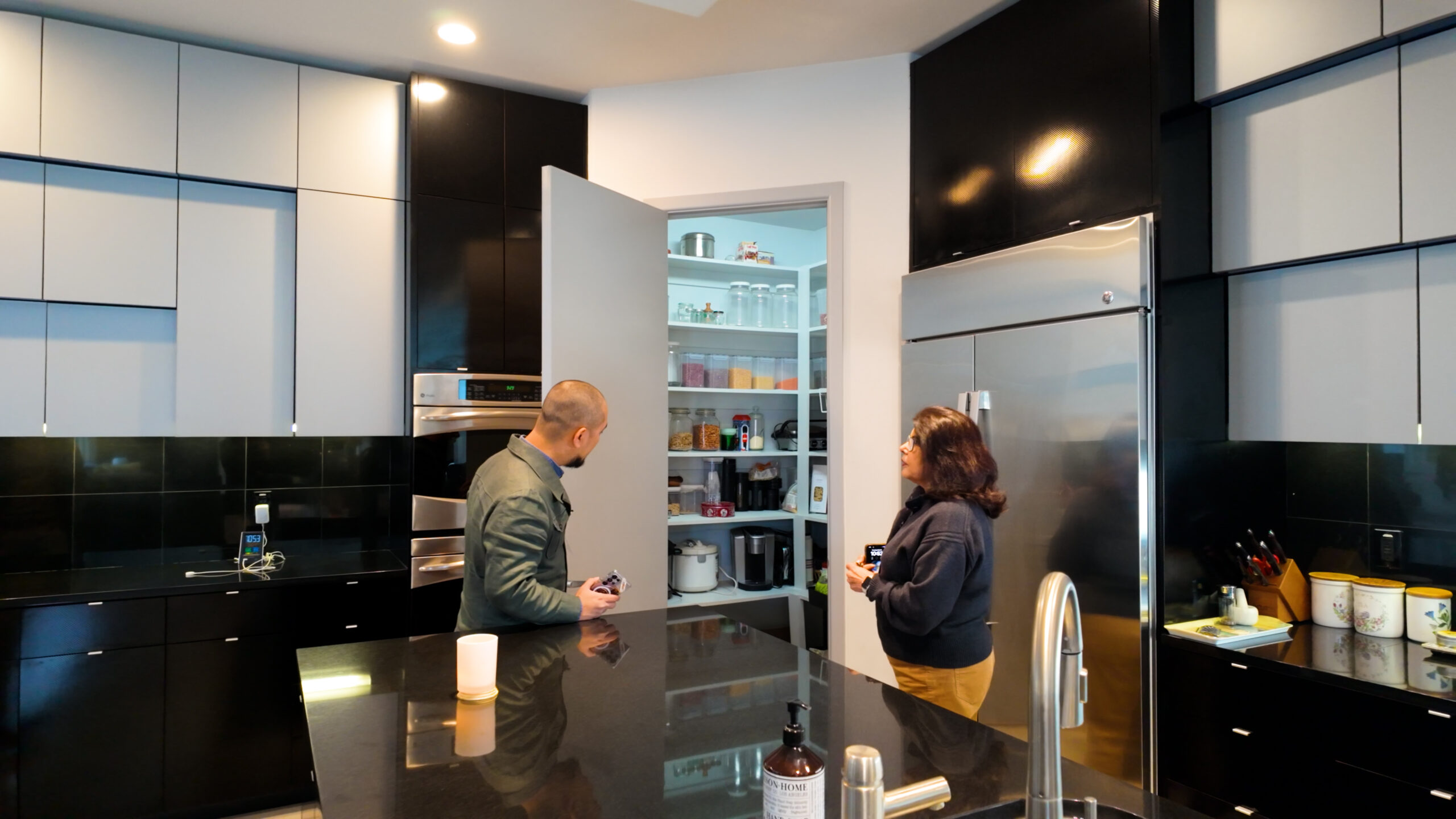 6-2-scaled-1-1 Nhà bếp hiện đại với hai người trò chuyện trước cửa tủ thực phẩm mở, căn bếp có thiết kế tối giản và sáng sủa.