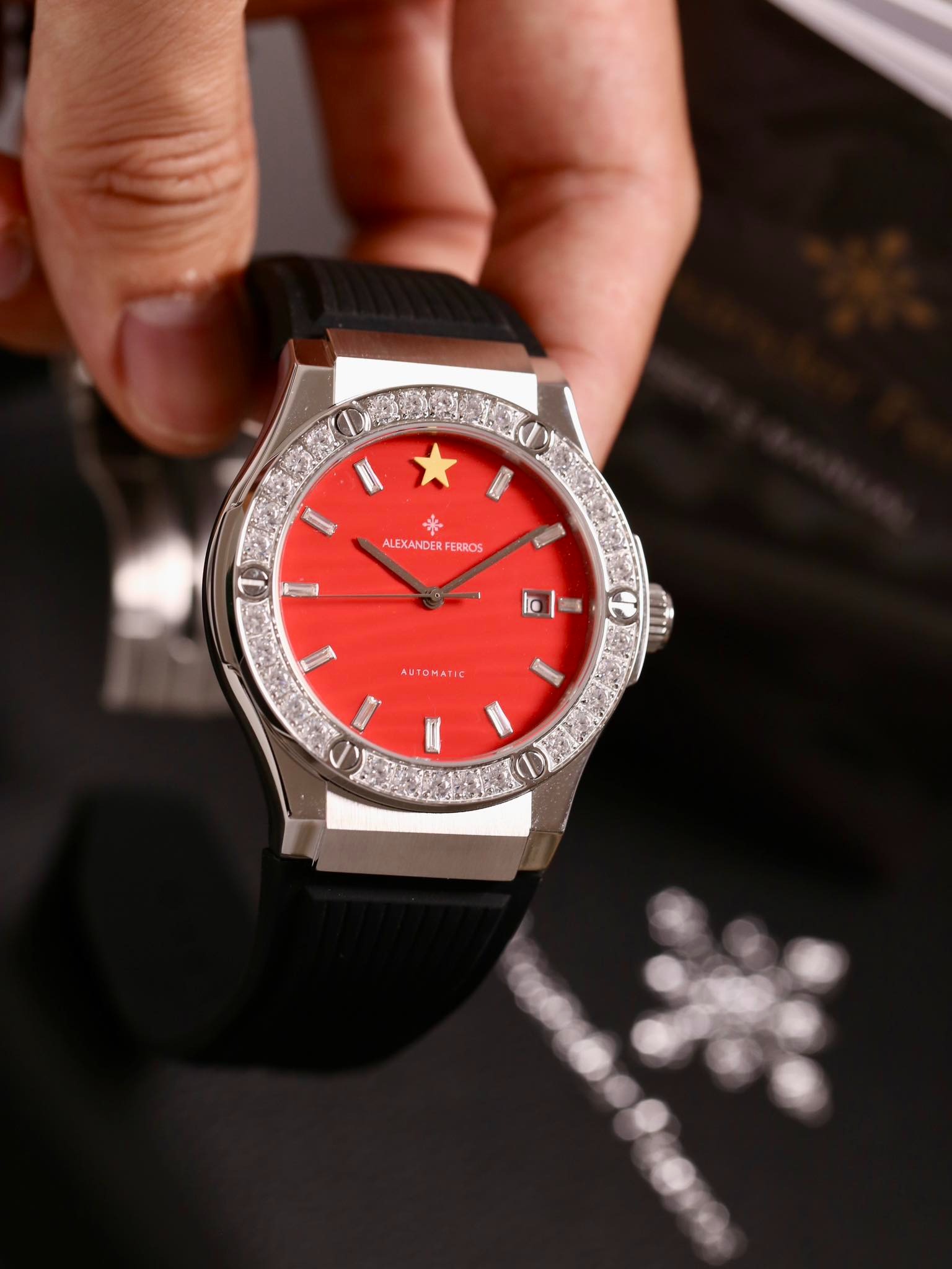 Đồng hồ nam mặt đỏ, viền đá quý, dây đen, được cầm trên tay tạo phong cách sang trọng và tinh tế.