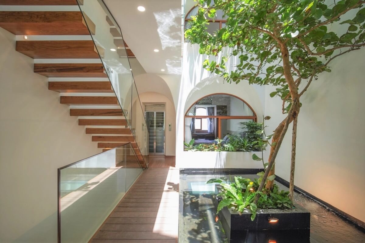 Cầu thang gỗ hiện đại với lan can kính bên cạnh vườn cây trong nhà, không gian sáng sủa và thoáng đãng.