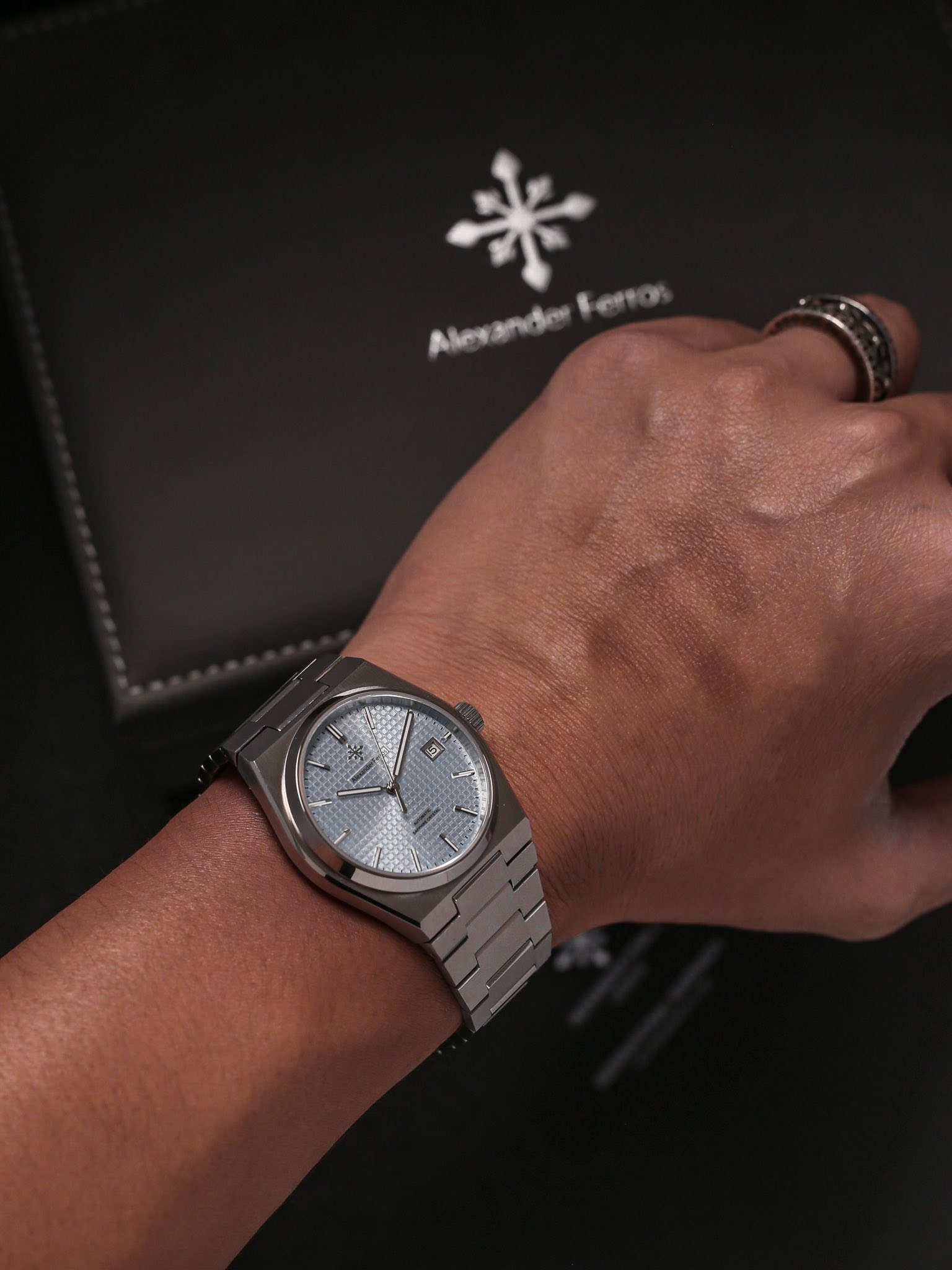 Đồng hồ đeo tay kim loại màu bạc trên tay người với hộp thương hiệu Alexander Ferros phía sau.