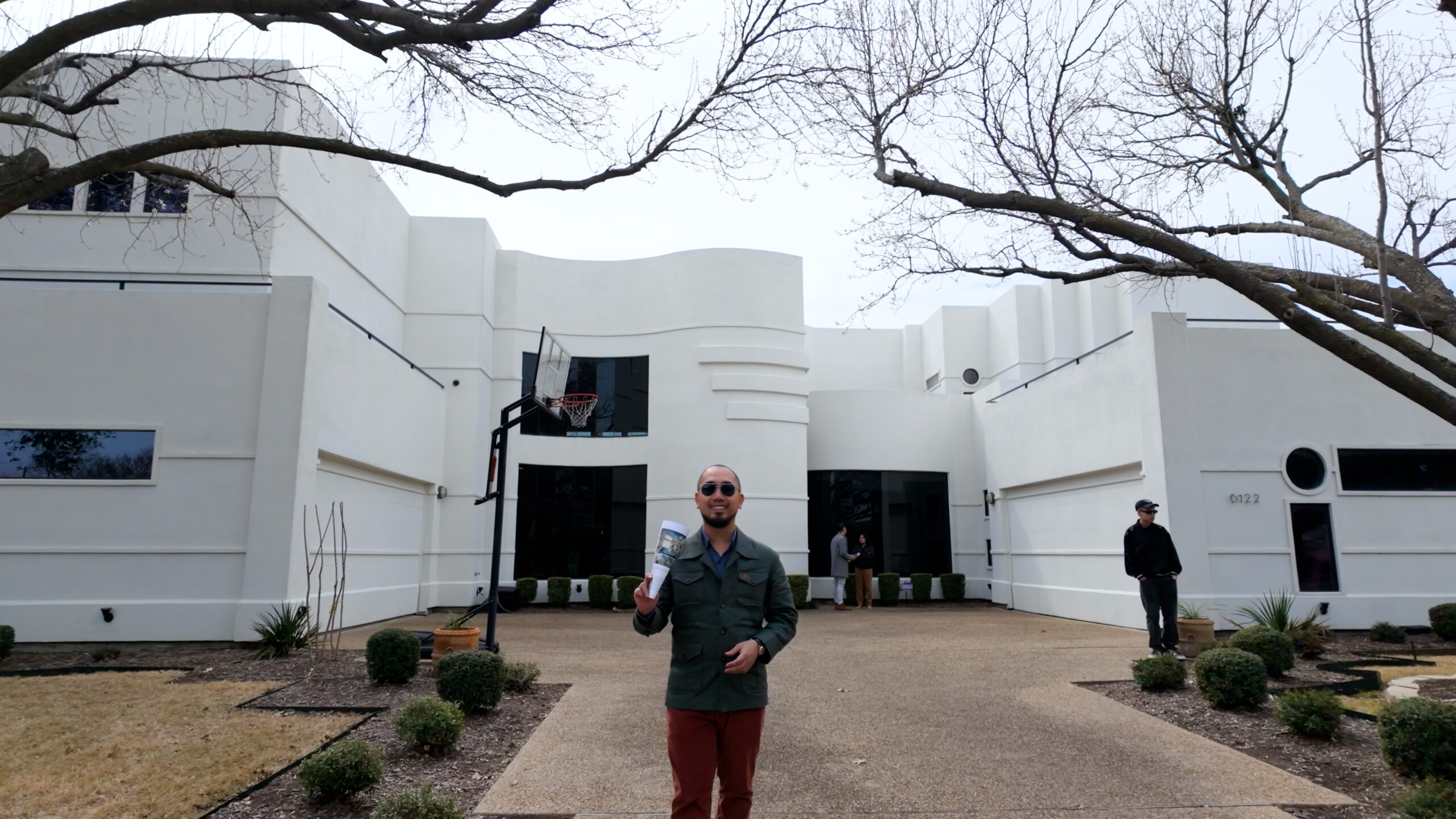 Người đàn ông đứng trước biệt thự hiện đại trắng, cầm bản vẽ thiết kế, cây khô phía trên.