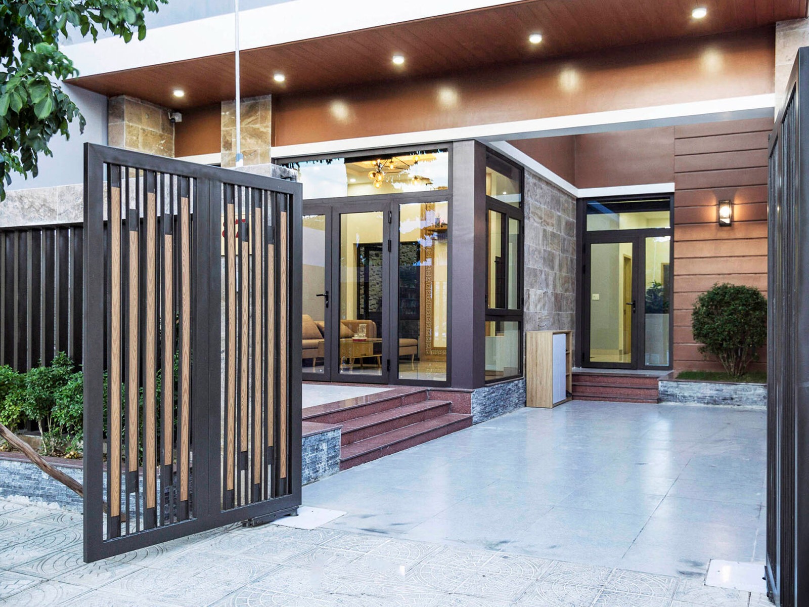 Lối vào ngôi nhà hiện đại với cửa kính và sân lát gạch, thiết kế tối giản, cây xanh bên cạnh cổng sắt.
