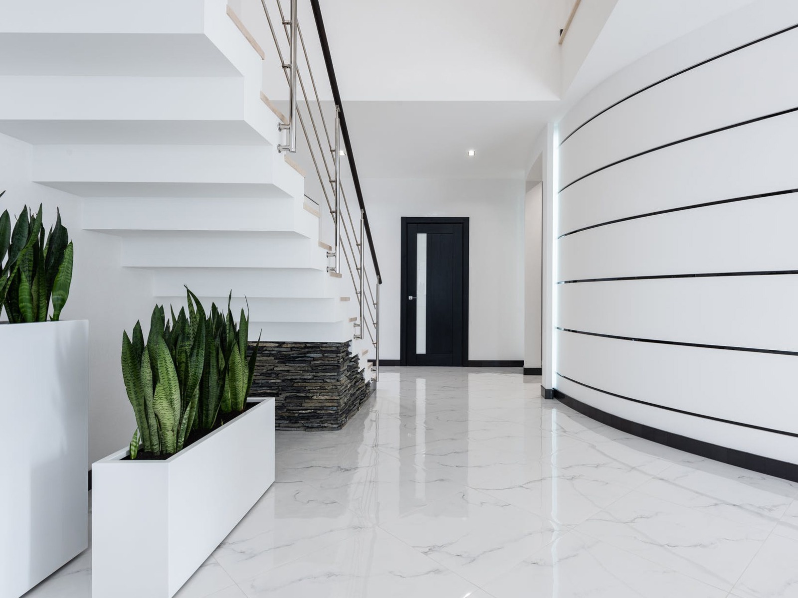 Cầu thang hiện đại với chậu cây xanh, sàn gạch trắng, tường cong trang trí đen, cửa gỗ màu tối.