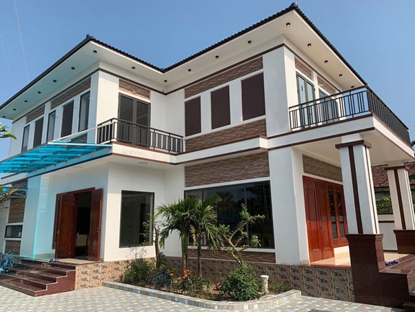 Nhà hai tầng hiện đại với mái ngói xanh, cửa kính rộng, và sân vườn nhỏ.
