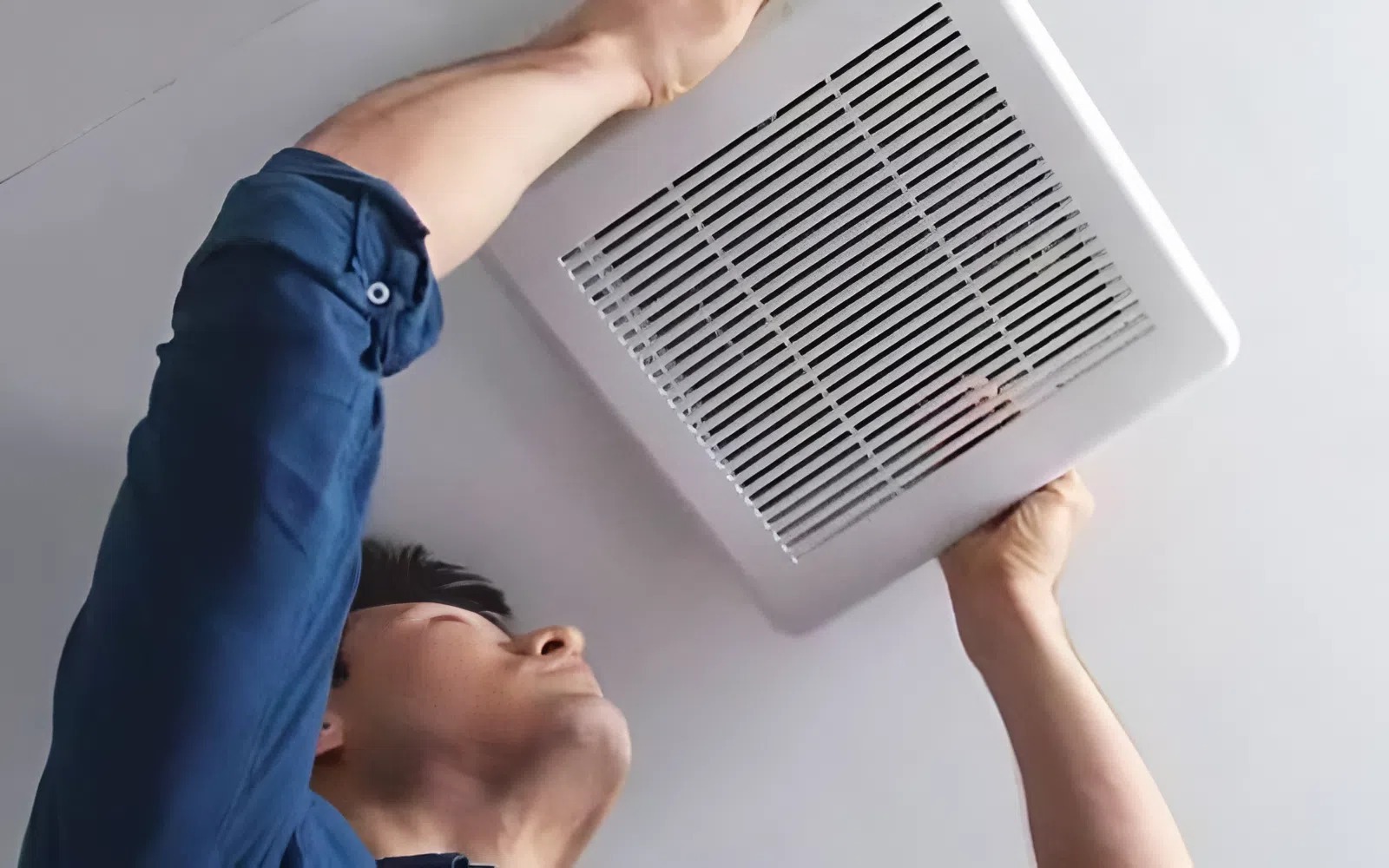 Người thợ lắp đặt quạt thông gió trần màu trắng trong phòng, mặc áo sơ mi xanh.