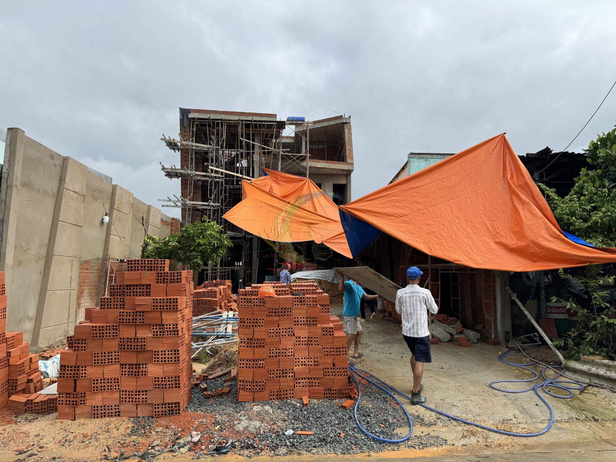 Công trình xây dựng nhà ở với vật liệu gạch đỏ và bạt che nắng màu cam, hai công nhân đang làm việc.