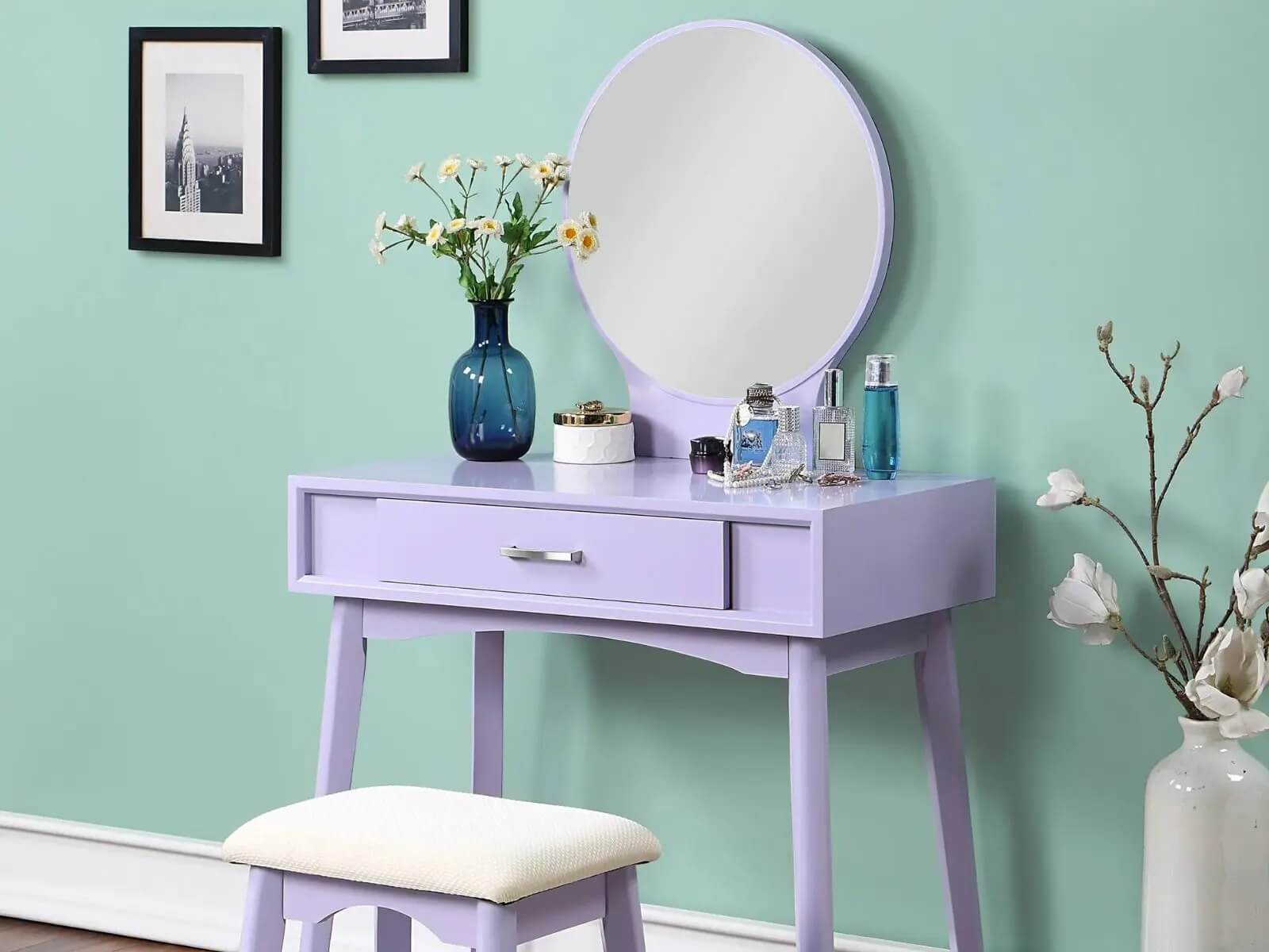 Bàn trang điểm màu tím nhạt với gương tròn, bình hoa xanh, và ghế đệm trắng, nổi bật trên nền tường xanh ngọc.