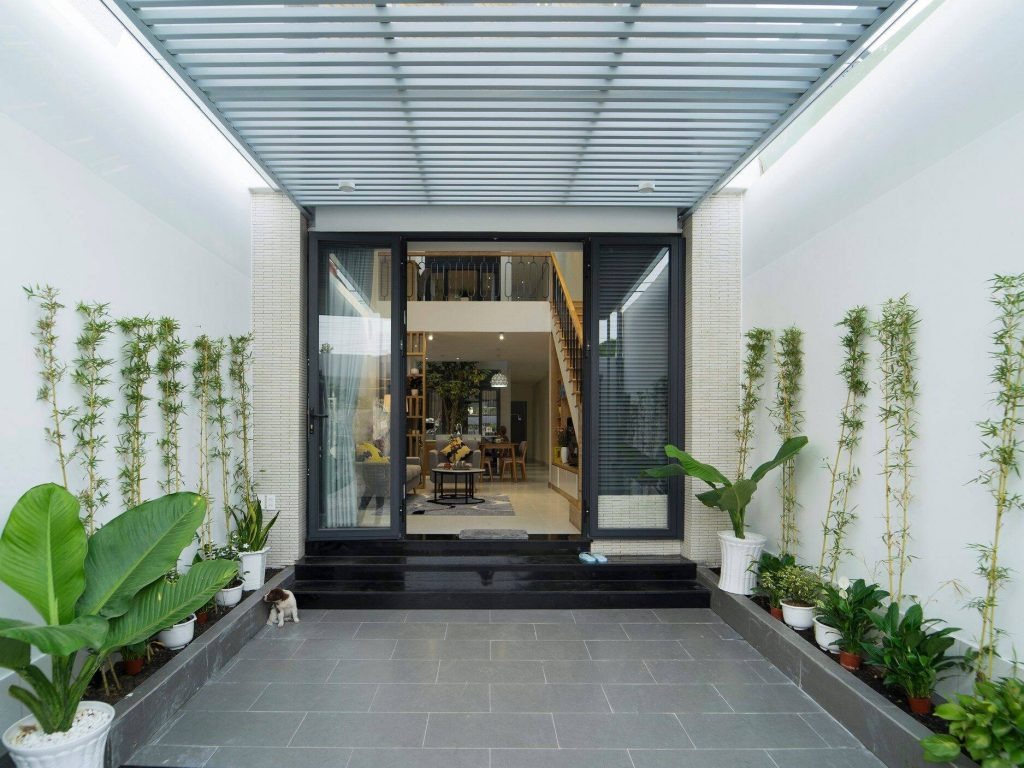 Lối vào nhà hiện đại với nhiều cây cảnh, cửa kính lớn và mái che sáng tạo.