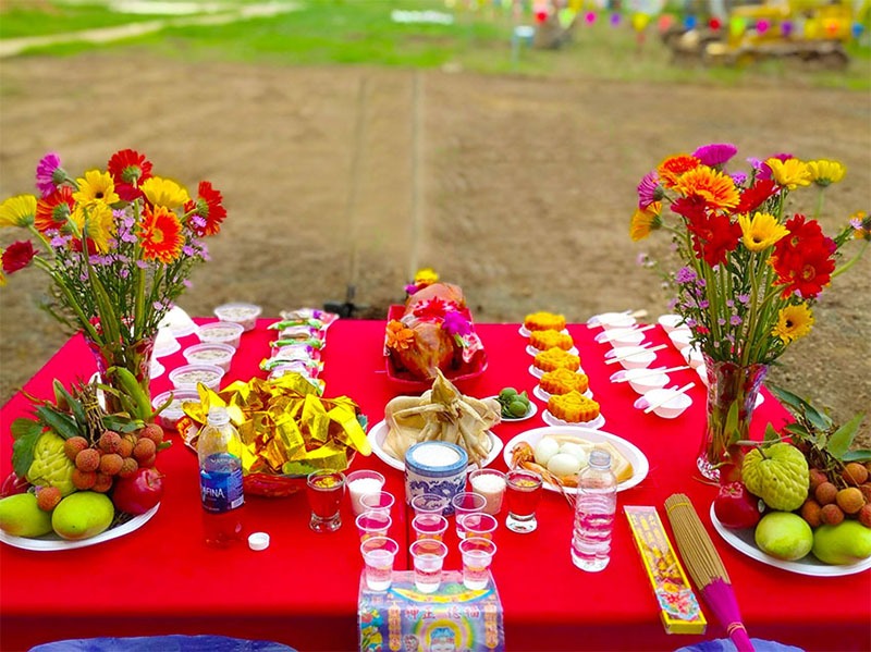 Bàn cúng lễ với hoa, trái cây và gà luộc trên khăn đỏ trong không gian ngoài trời.