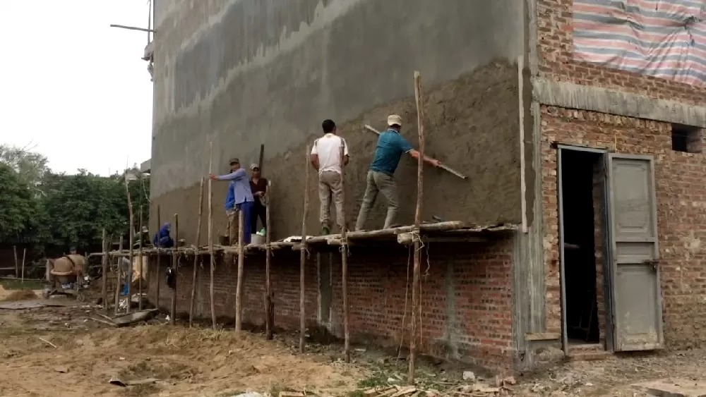 Công nhân xây dựng trát vữa lên tường gạch, đứng trên giàn giáo gỗ tại công trình xây dựng.