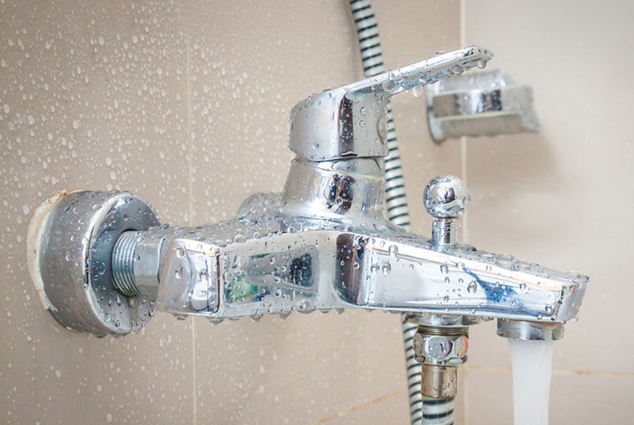 Vòi sen kim loại màu bạc với nước đang chảy trong phòng tắm có gạch màu be.