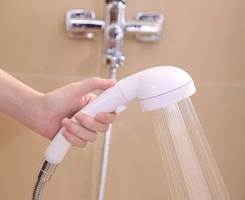 cach-mo-voi-sen-tam-thuong Tay cầm vòi sen trắng đang chảy nước trong nhà tắm, lắp đặt trên gạch màu be.