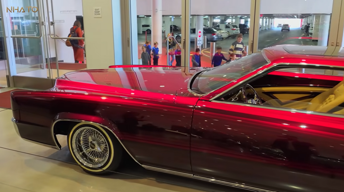 cadillac-do Xe cổ màu đỏ bóng loáng trưng bày trong showroom với nhiều người xem qua cửa kính phía sau.