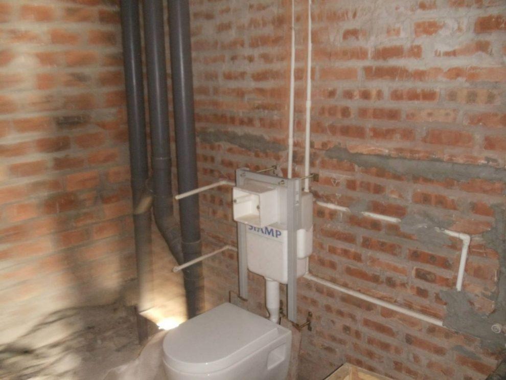 Nhà vệ sinh đang xây, tường gạch đỏ lộ, ống dẫn và hệ thống nước chưa hoàn thiện.