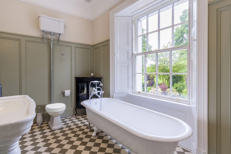 Phòng tắm cổ điển với bồn tắm đứng cạnh cửa sổ lớn, sàn gạch caro và nhà vệ sinh kiểu cũ.