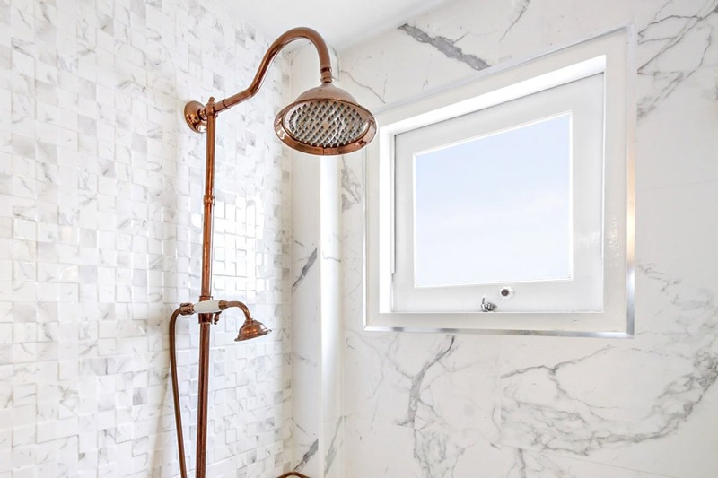 Vòi sen đồng trong phòng tắm ốp đá cẩm thạch trắng sang trọng, cạnh cửa sổ kính lớn.