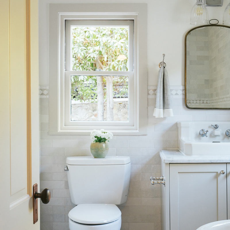 Phòng tắm sáng sủa với bồn cầu trắng, cửa sổ lớn, cây xanh bên ngoài và bồn rửa tay có gương.