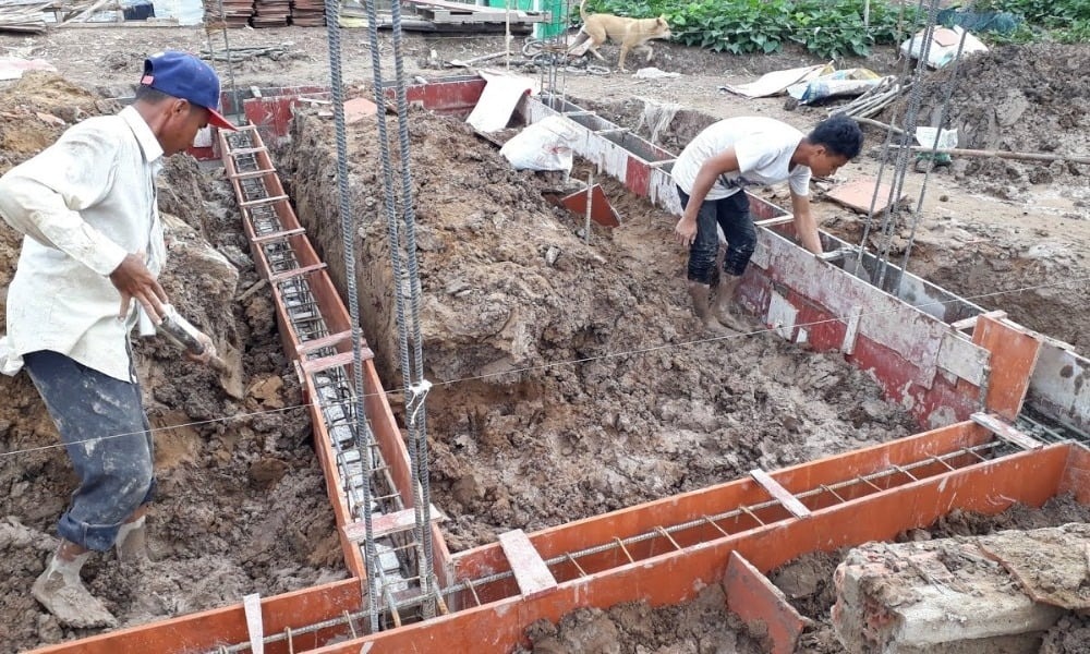 Công nhân xây dựng làm việc trên công trường nền móng bê tông, có đất và vật liệu xây dựng xung quanh.