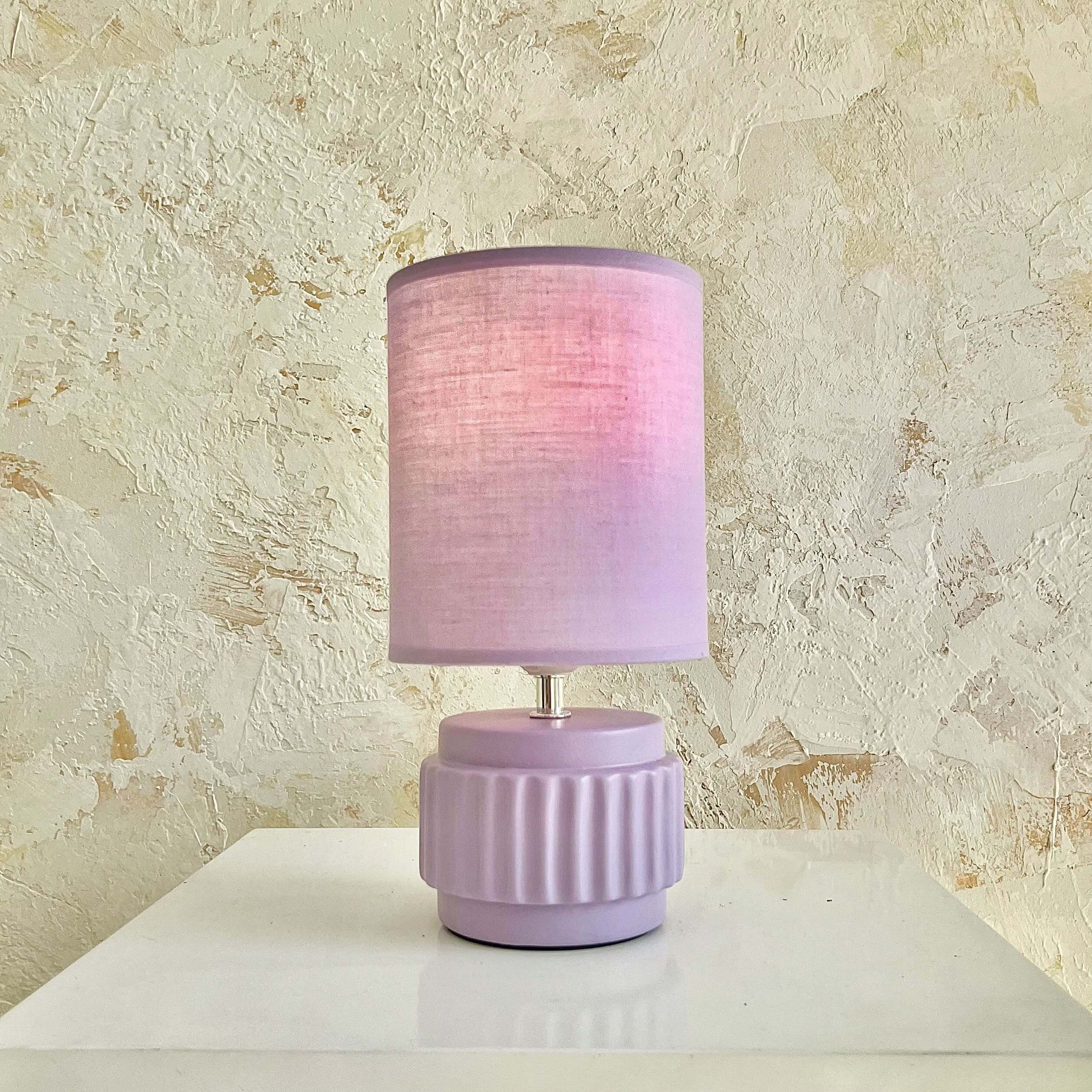 Đèn bàn màu tím nhạt trên mặt bàn trắng nền tường vân nổi, tạo không gian ấm cúng và phong cách.