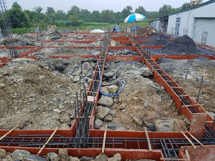 do-mong-nha-ngay-mua-khien-giam-chat-luong-cua-vat-lieu-xay-dung Công trình xây dựng móng nhà với thép gia cố và nền đất đang thi công, có ô che nắng màu xanh trắng.