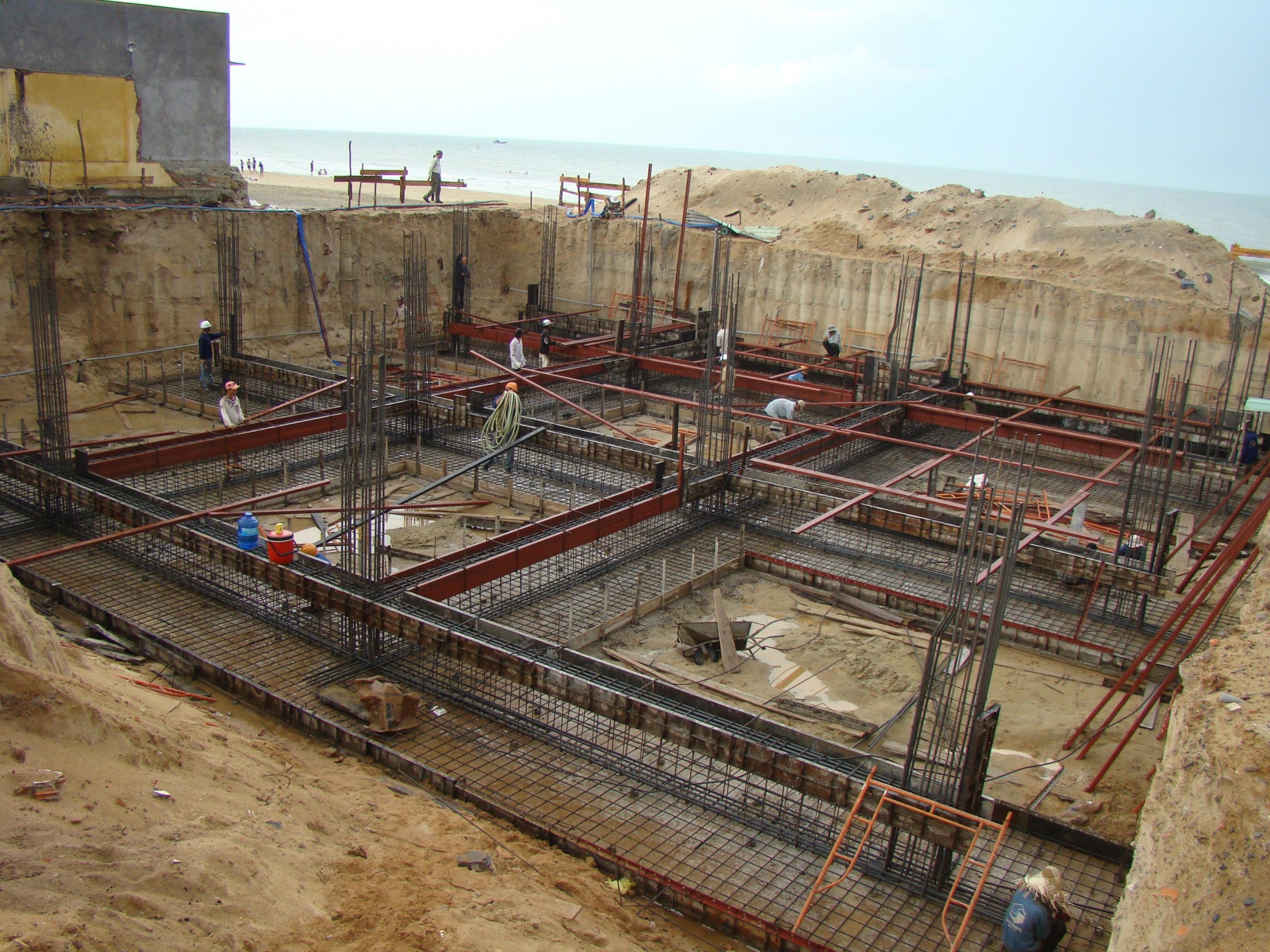 Công trình xây dựng móng bê tông với công nhân đang làm việc gần bãi biển, khung thép và sàn cát.