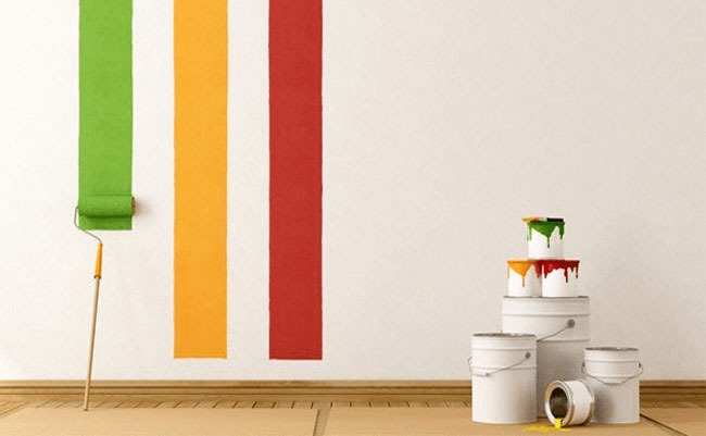 Con lăn sơn trên tường với các dải màu xanh, vàng, đỏ và thùng sơn xếp chồng, nền gỗ nhạt, ánh sáng tự nhiên.