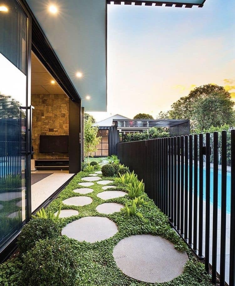 Lối đi sân vườn lát đá tròn, bên cạnh là hàng rào và hồ bơi, cạnh hiên nhà hiện đại.