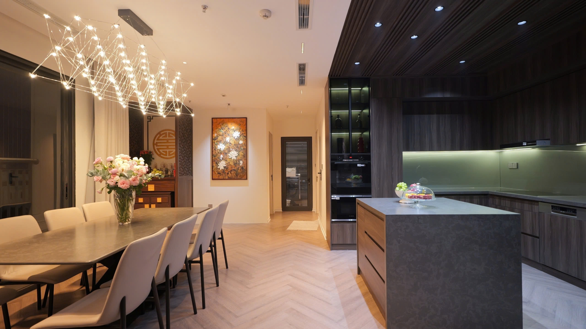 dz0pfcm6t15qroi92bvh Nhà bếp hiện đại với bàn ăn lớn, đèn treo, bức tranh nghệ thuật và tủ gỗ tối màu.
