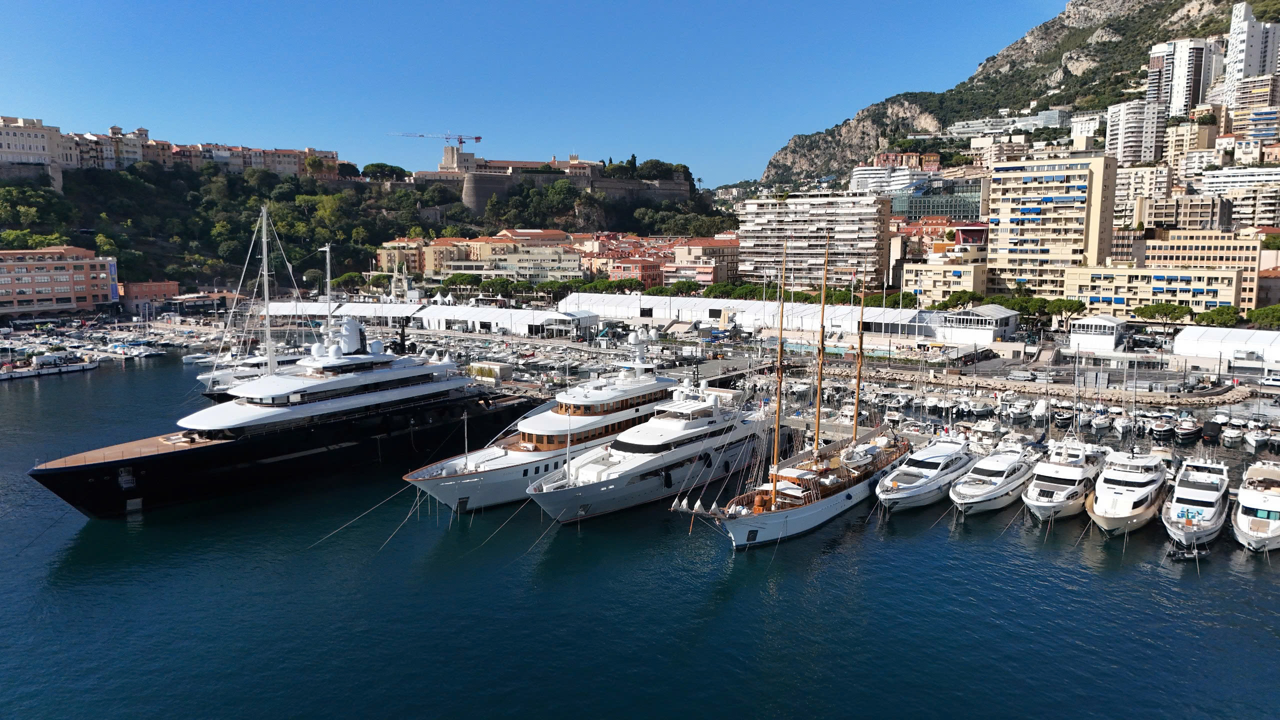 Du thuyền sang trọng neo đậu tại bến cảng Monaco dưới ánh nắng rực rỡ, phía sau là cảnh quan đô thị.