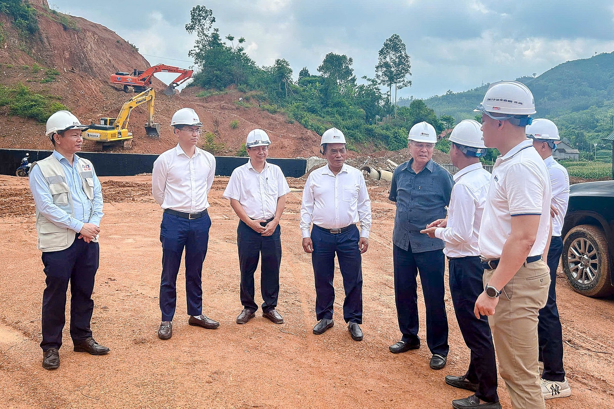 giai-phap-cho-viec-khoi-cong-trong-mua Nhóm kỹ sư đội mũ bảo hộ thảo luận tại công trường với máy xúc đất hoạt động phía sau.