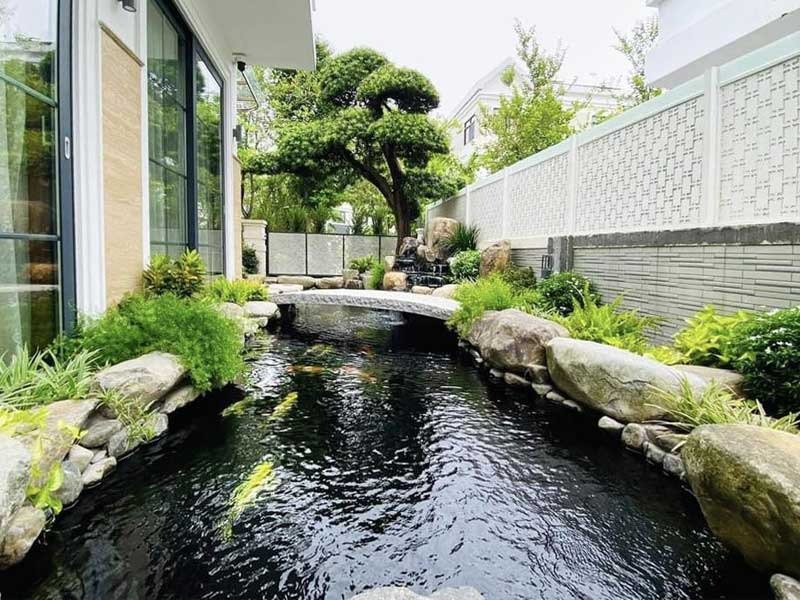 Hồ cá koi trong vườn với cầu đá, cây xanh tươi tốt tạo không gian yên bình, thư giãn.