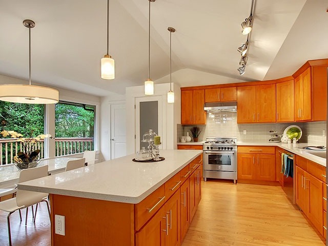 Nhà bếp hiện đại với tủ gỗ, bàn đá, đèn treo, và cửa sổ nhìn ra ngoài trời xanh.