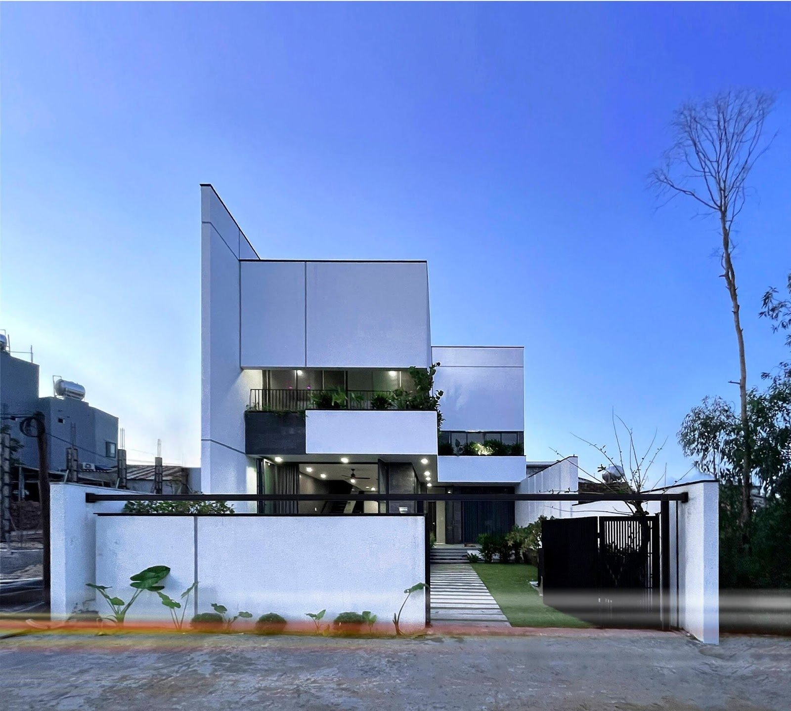 Ngôi nhà hiện đại ba tầng màu trắng với sân vườn, cây xanh xung quanh và bầu trời xanh trong cảnh hoàng hôn.