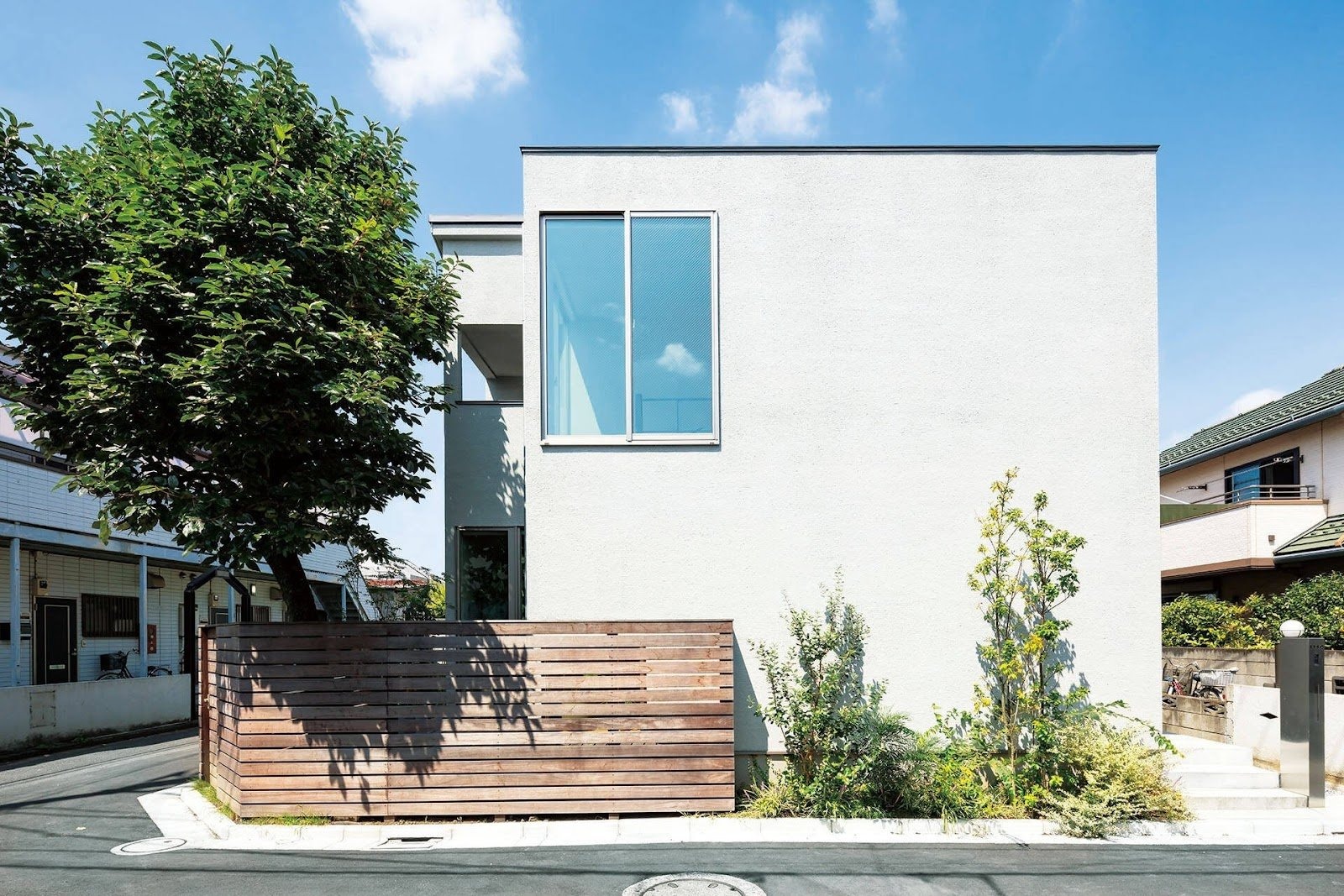 Ngôi nhà hiện đại với tường trắng, cửa sổ lớn, bên cạnh cây xanh và hàng rào gỗ, bầu trời trong xanh.
