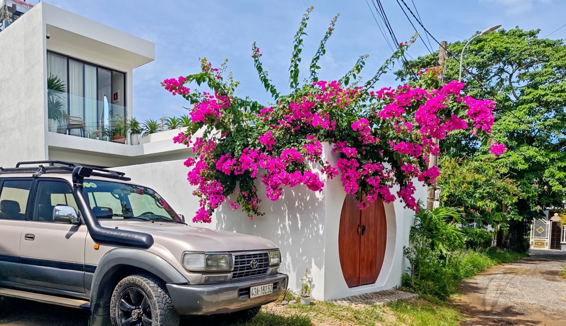 Ngôi nhà hiện đại với hoa giấy màu hồng rực rỡ, xe ô tô đỗ phía trước, bầu trời trong xanh.
