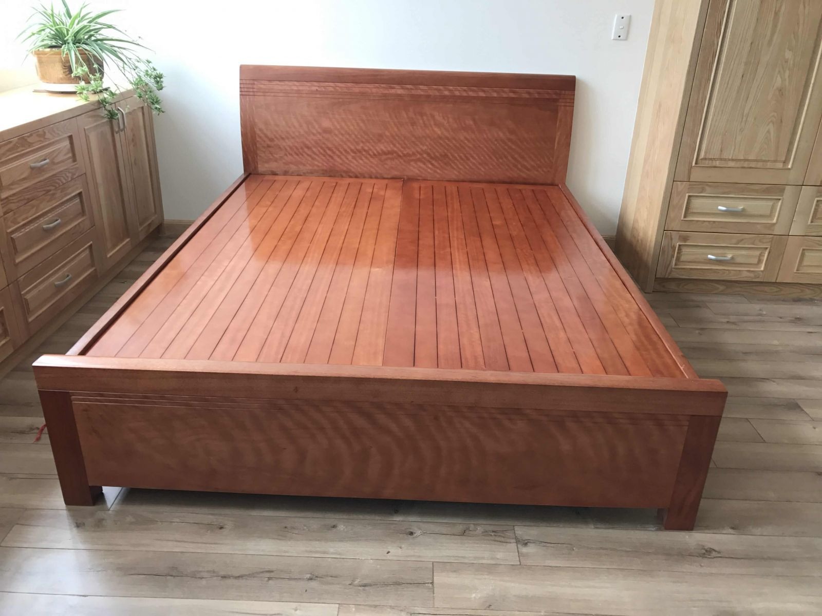 Giường gỗ nâu sáng truyền thống trong phòng với tủ và cây xanh trang trí bên cạnh.