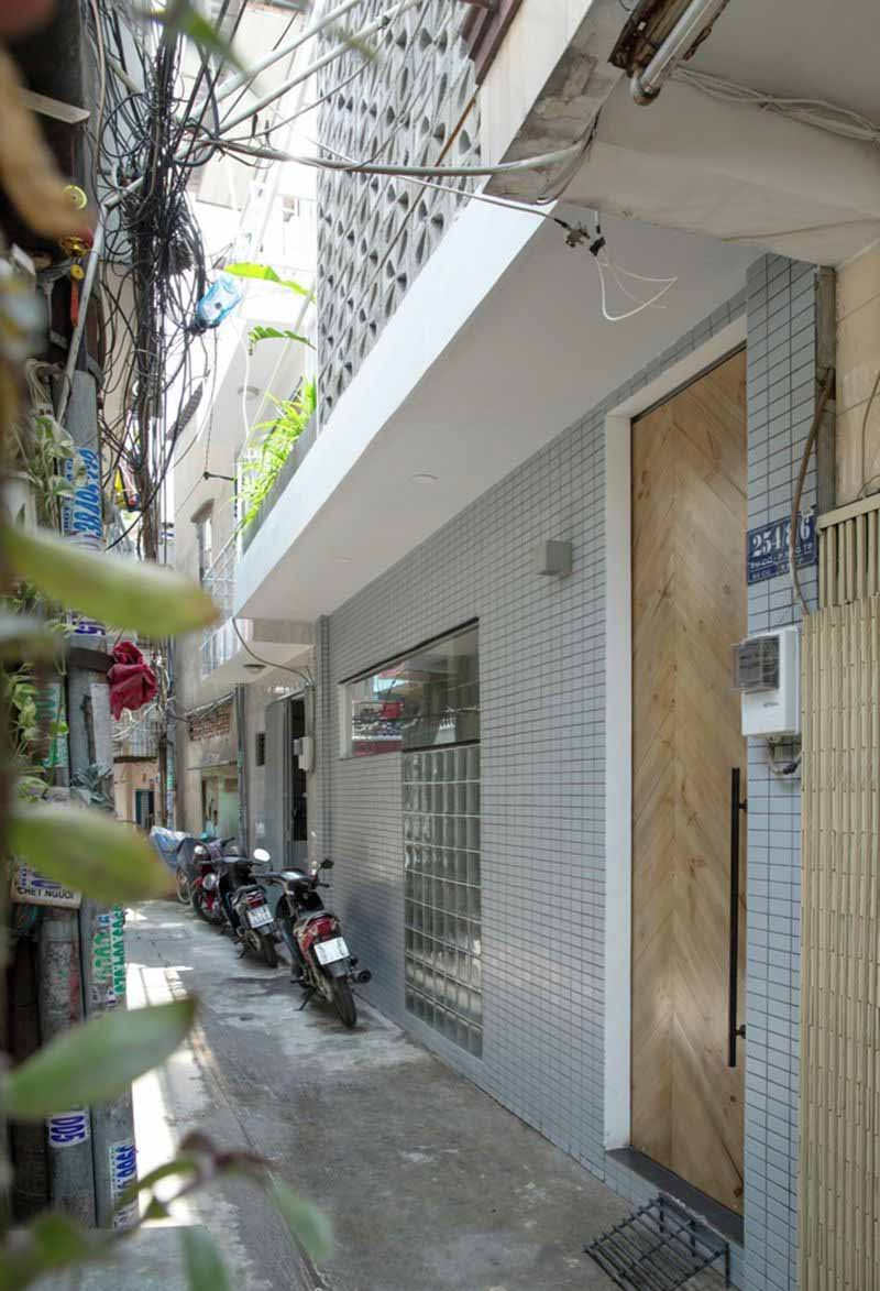 Ngõ hẹp với xe máy đậu, bên cạnh là tường ốp gạch và cửa gỗ hiện đại. Cáp điện chạy dọc theo đường.