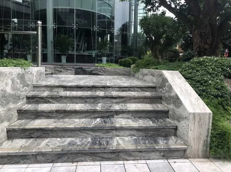 Cầu thang đá cẩm thạch trước tòa nhà kính hiện đại, xung quanh có cây xanh trang trí.