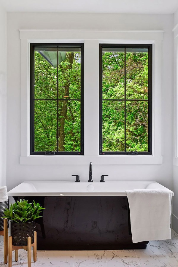 Bồn tắm trắng bên cửa sổ lớn nhìn ra cảnh rừng xanh tươi, trang trí thêm chậu cây và khăn tắm mềm mại.