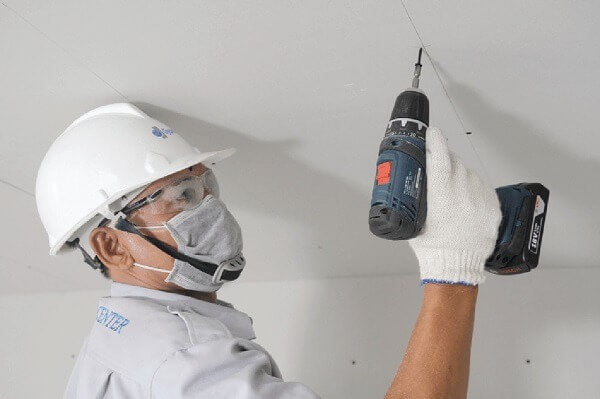 ky-thuat-lap-dat-tran-thach-cao-chim-chinh-xac-3 Kỹ sư xây dựng đang khoan trần nhà, đội mũ bảo hộ và đeo khẩu trang an toàn.