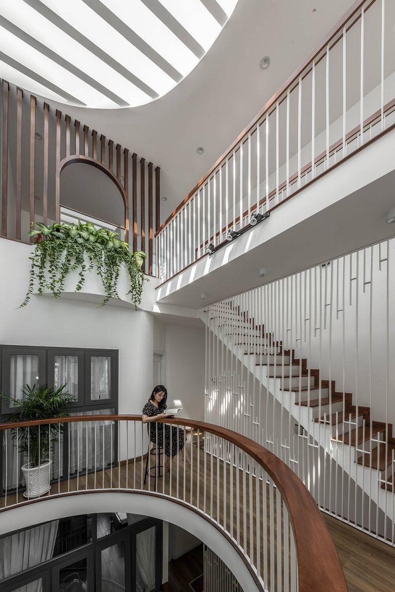 Nhà hiện đại với cầu thang nổi bật, ánh sáng tự nhiên xuyên trần, cây xanh trang trí, và người phụ nữ đọc sách.