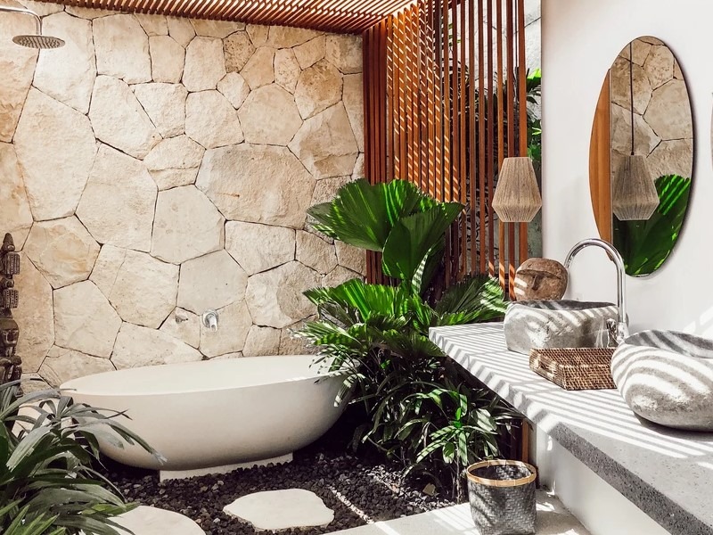 Phòng tắm ngoài trời phong cách tự nhiên, bồn tắm trắng, tường đá, cây xanh và gương trang trí.