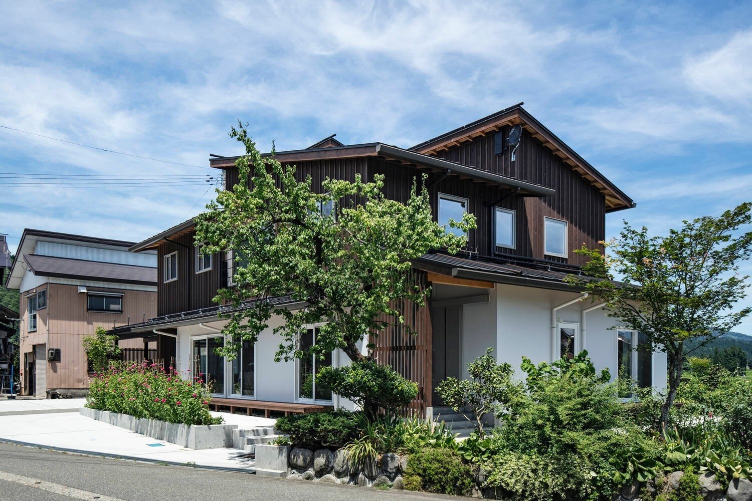 Ngôi nhà gỗ hai tầng kiểu truyền thống Nhật Bản với cây xanh, kèm khung cảnh trời xanh và mây trắng.